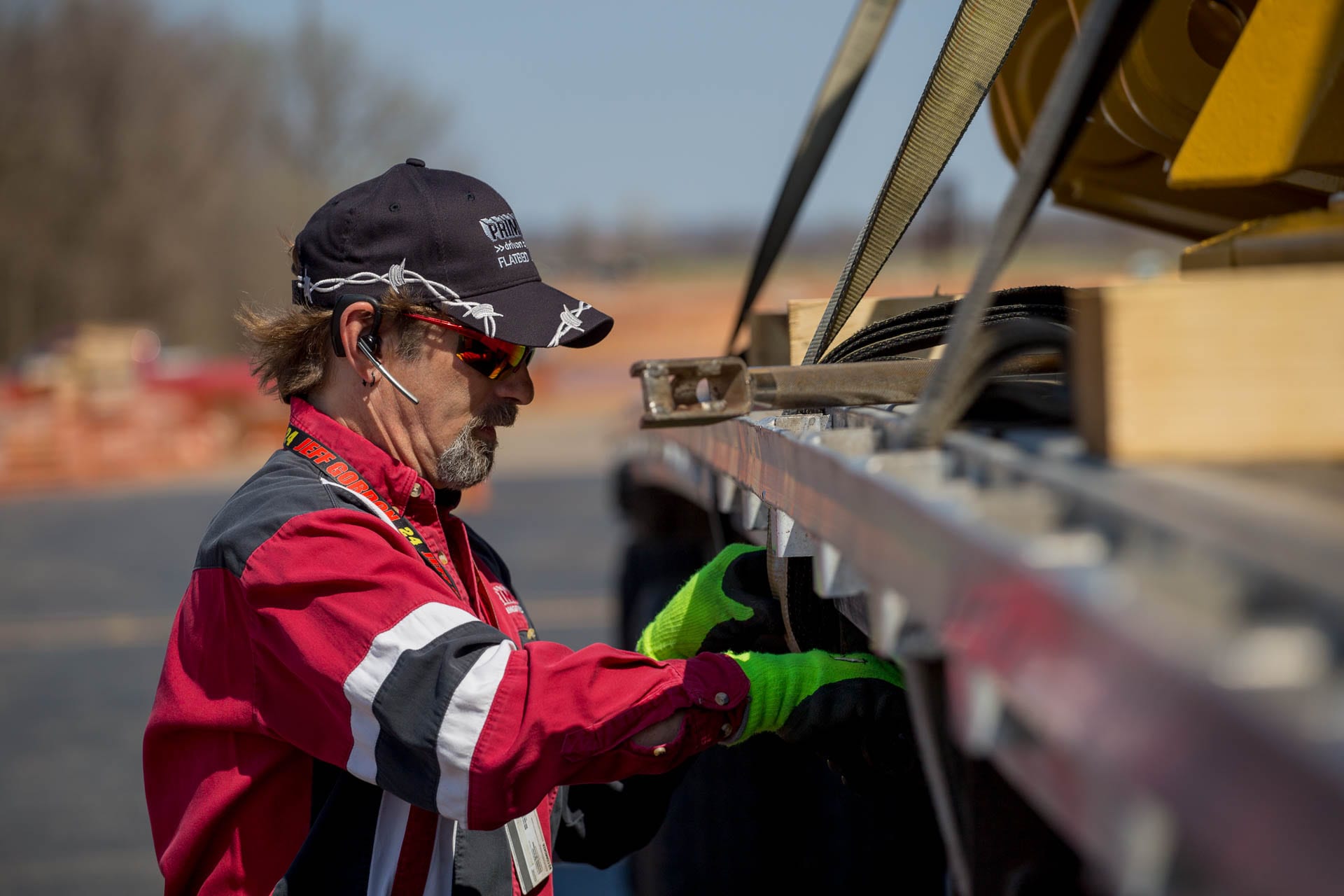 A Prime truck driver. wearing a black hat & red jacket, tying down a load on a flatbed trailer.