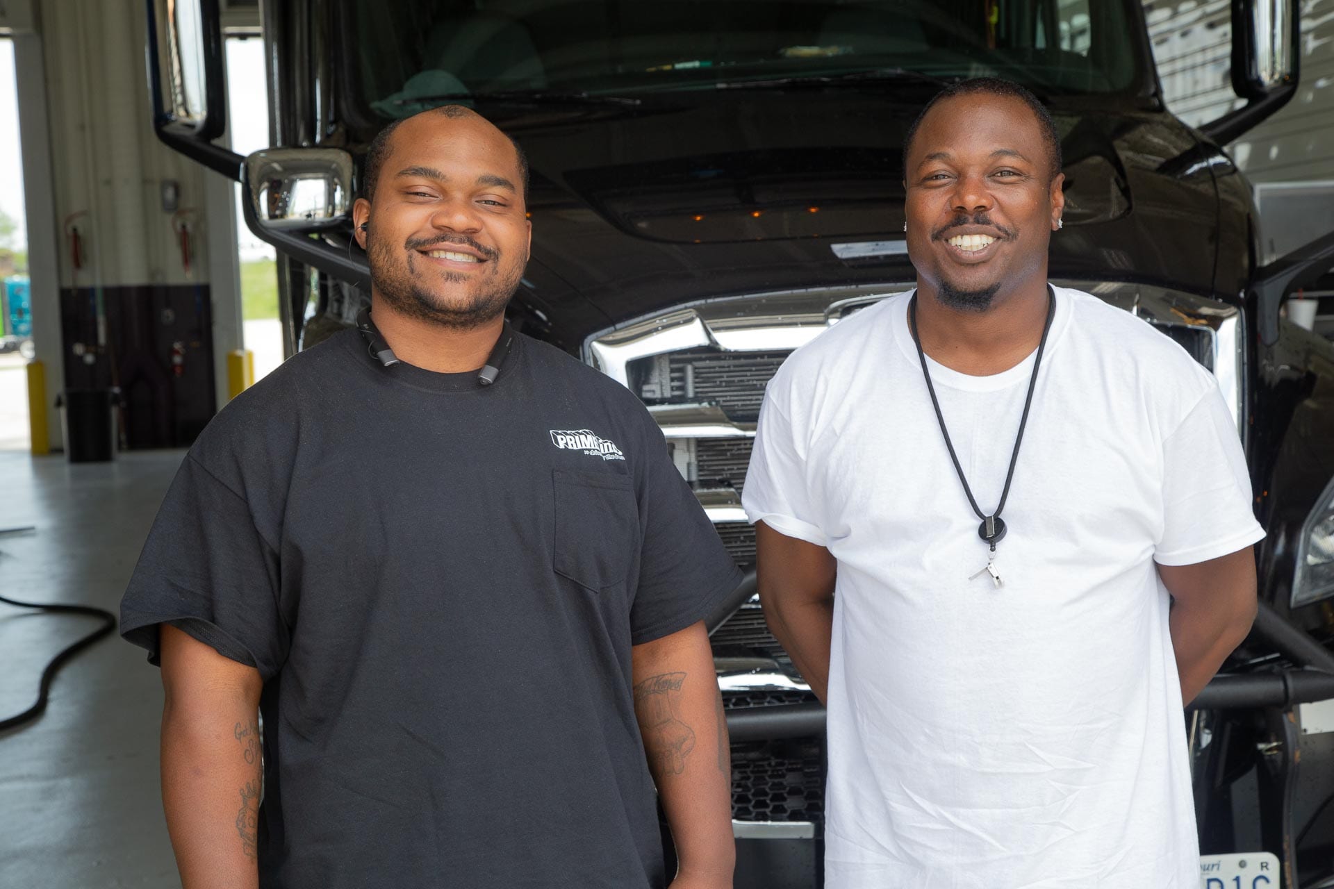 Two Prime truck drivers posing for a picture in front of a black Freightliner semi-truck.