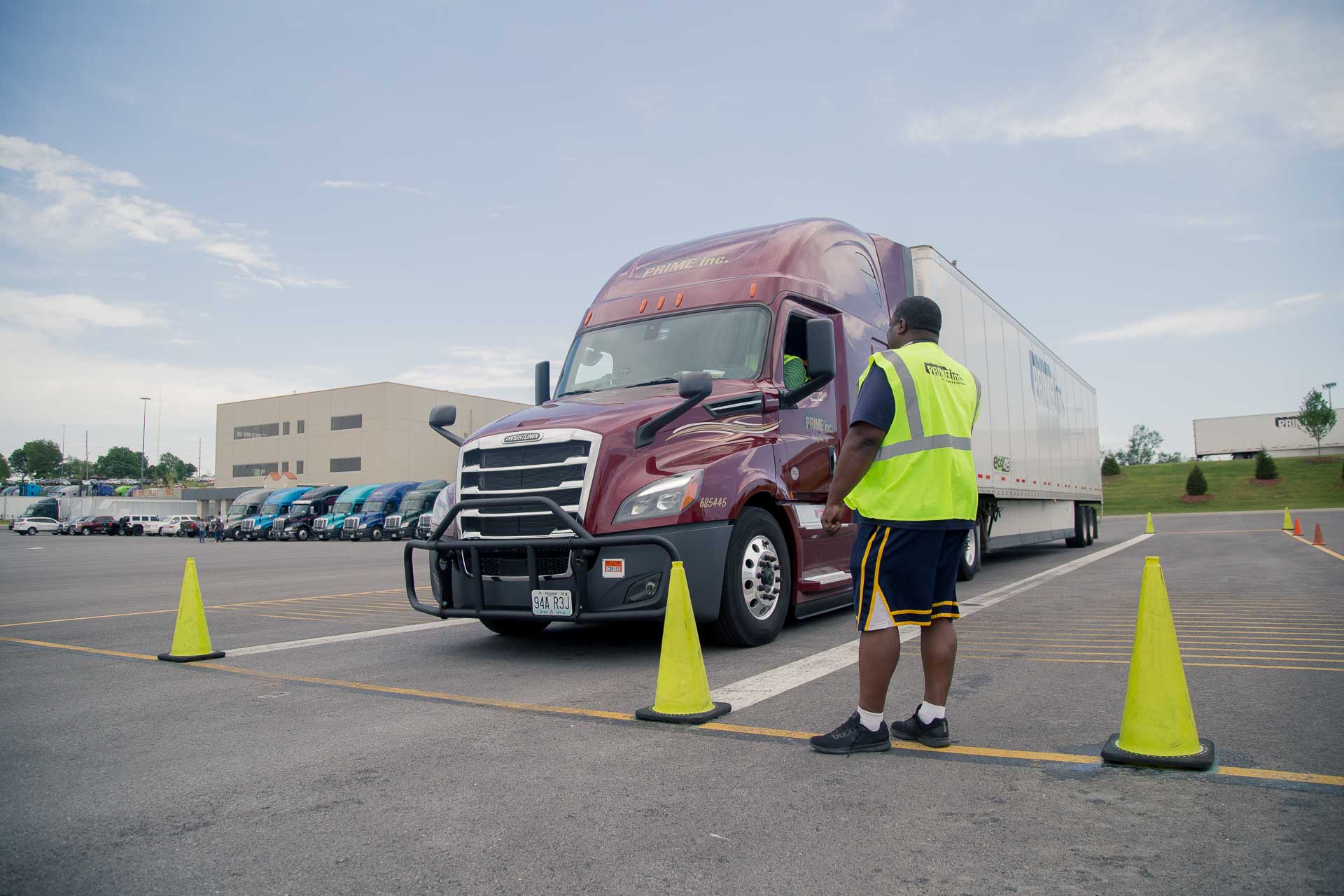 A Prime driving instructor observing a student driver in a red semi-truck driving in a parking lot.