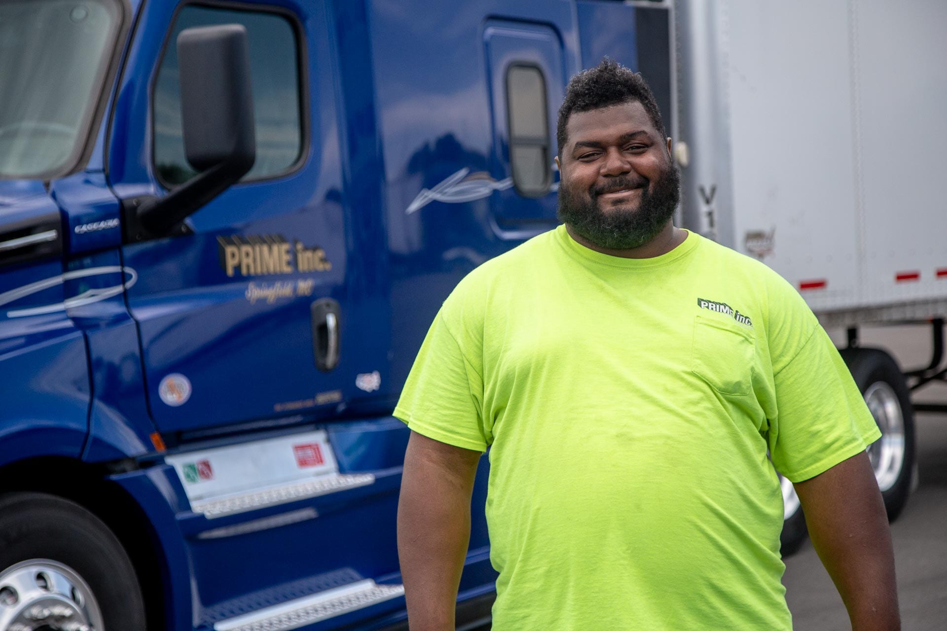 A Prime truck driver, wearing a safety yellow t-shirt, standing in front of a dark blue semi-truck.