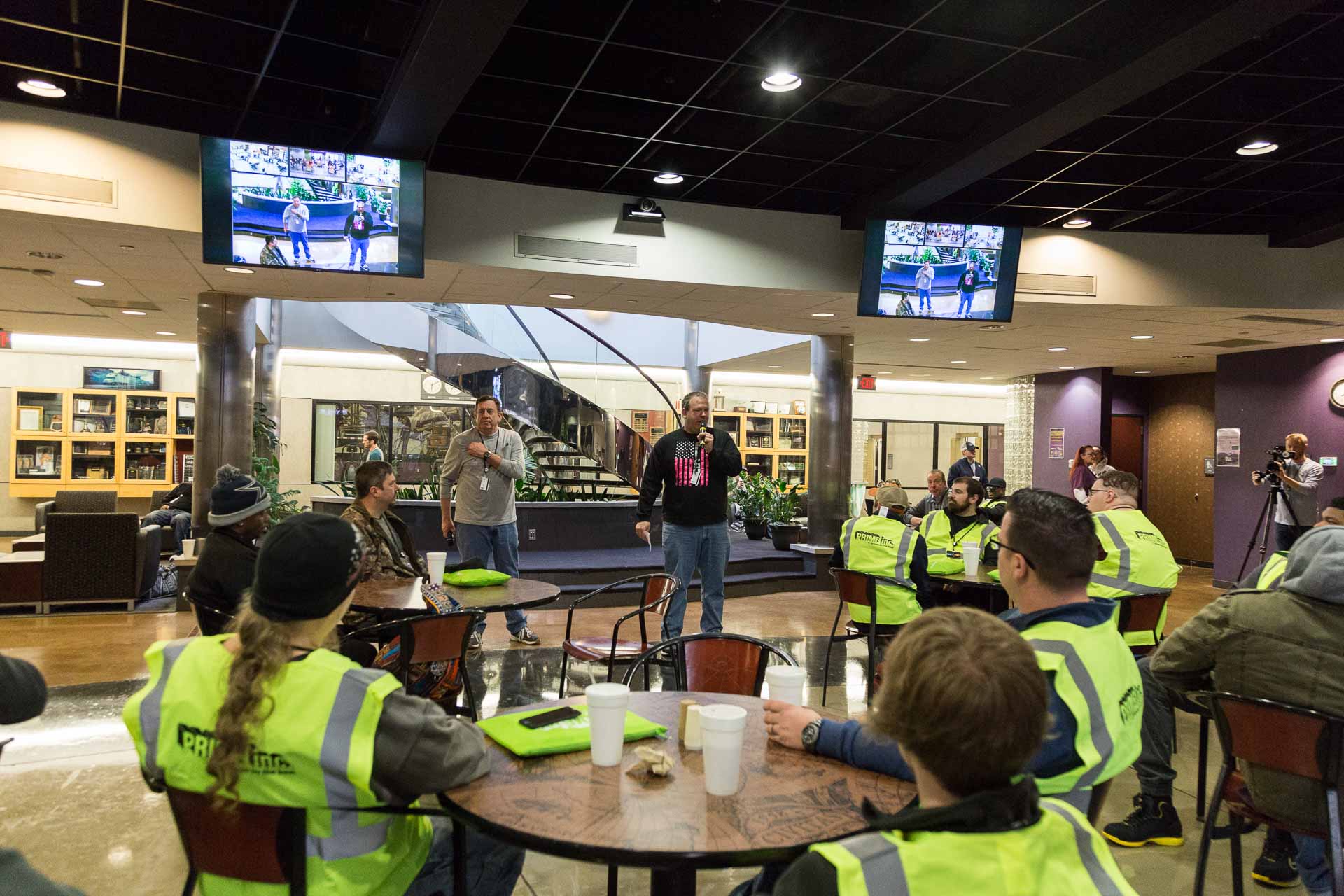 A Prime Inc. associate speaking to his team in a cafeteria area who are all wearing bright green safety vests.