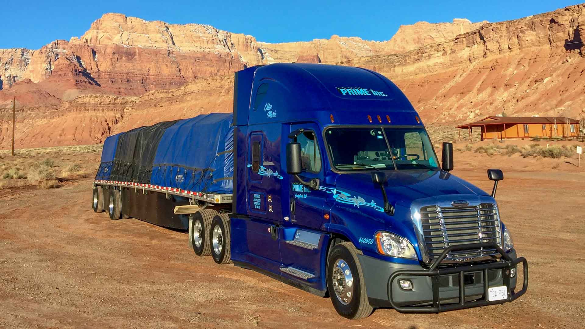 Blue Freightliner semi-truck towing a covered flatbed trailer surrounded by a rocky landscape.