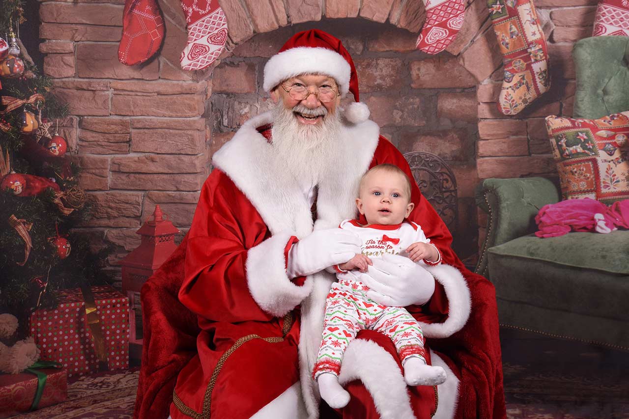 Santa Claus, taking a picture with a young child who is sitting on his lap dressed in a red and white christmas outfit at Prime, Inc.'s annual Santa Claus visit.