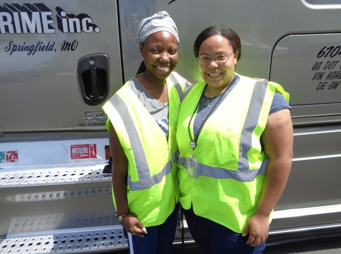 Two of Prime Inc's female truck drivers, standing in front of a brown semi-truck, wearing bright yellow safety vests.