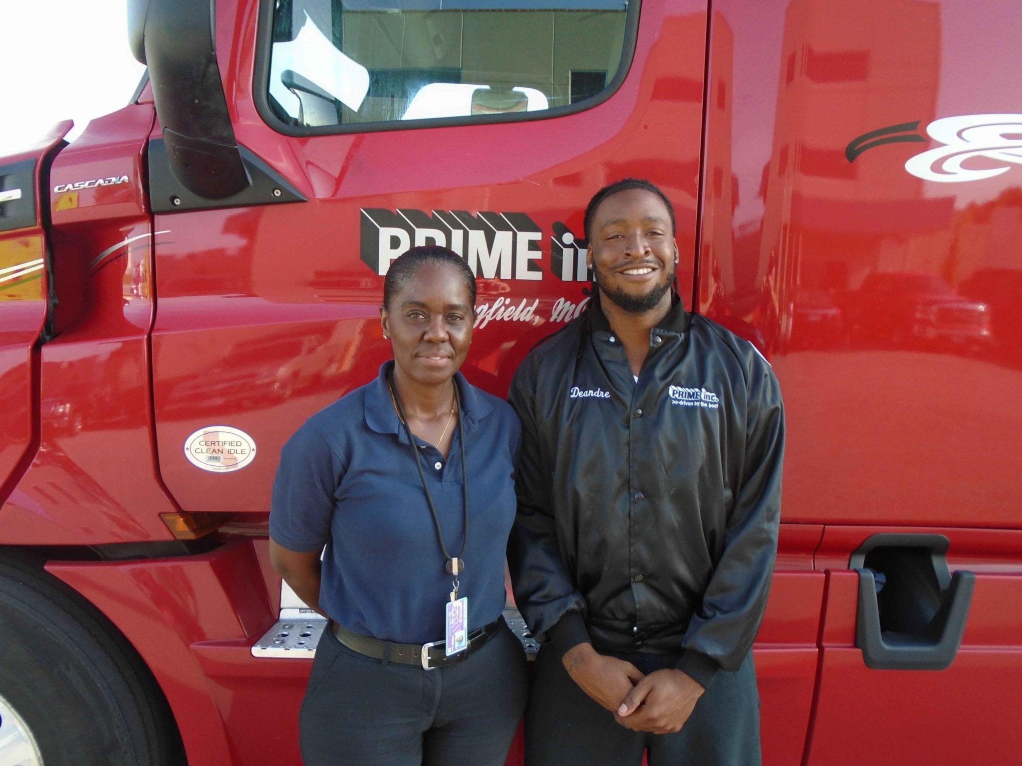Deandre, a Prime student truck driver. standing with his instructor in front of a red semi-truck.