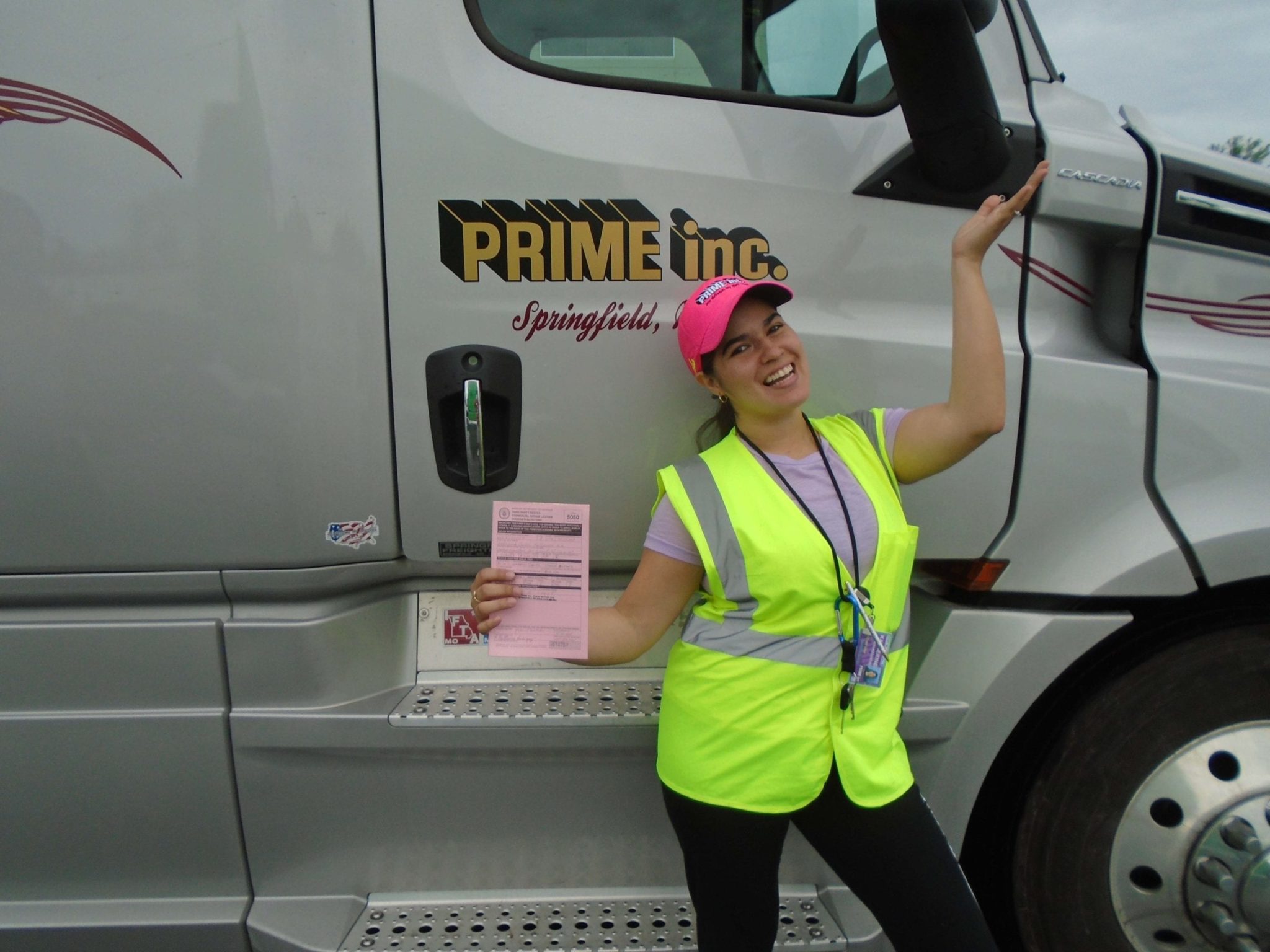 A Prime, Inc. female truck driver, wearing a bright safety vest and a pink hat, standing in front of a brown semi-truck while holding up her CDL exam scores.