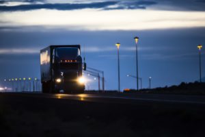 A semi truck drives on the highway at night with the backdrop of the setting sun.