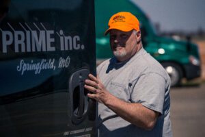 A man in an orange hat stops to look at the camera as he opens his semi-truck cab door to get in. The door of the truck says, "Prime Inc. Springfield, MO."
