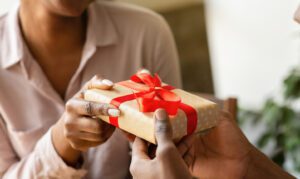 A person hands another person a small gift wrapped in tan paper with a red bow.