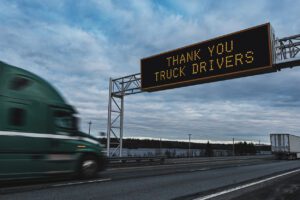 A highway sign reads "thank you truck drivers" as a semi-truck passes underneath.